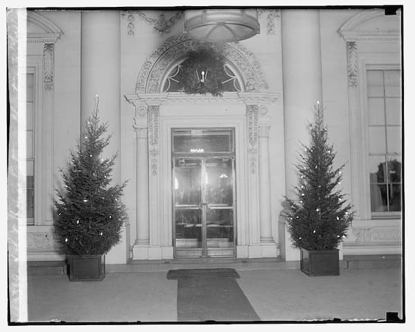 black and white photo of the entrance to the White House. Two Christmas trees with lights flank the glass doors