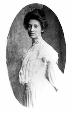 Mary Colter is dressed in a beautiful white dress trimmed with lace. The sleeves are long but puffy. Her hair is in a bouffant style and she is posing for the camera.