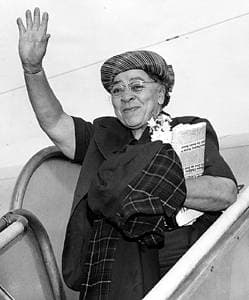 Charlotta Bass wears a jaunty hat and raises her right hand in greeting to the people meeting her on the airport tarmac. She carries a copy of her newsppaer in her left arm where she also holds a jacket or scarf.