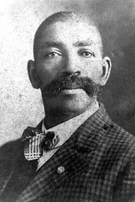 A professional photo of Bass Reeves. He is probably 35. His hair is very short, but he has a bushy handlebar moustache. He is in a jacket with a plaid bow tie.