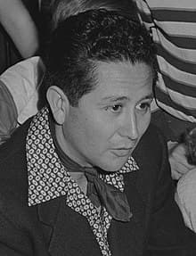 A black-and-white photograph of Gus Arriola. He has on a suit jacket, a printed shirt, and a scarf around his neck. 