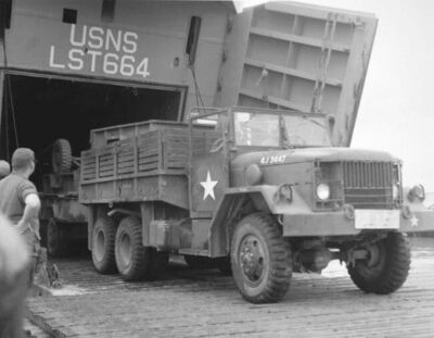 A GMC truck being unloaded from a boat as men watch