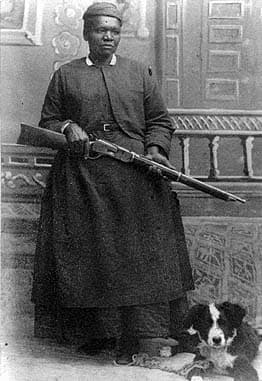 a black-and-white photograph of Mary Fields. She holds her rifle, and a black-and-white dog lies beside her.