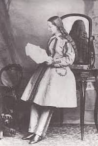 This is a girlhood photo of Mary Walker. Her hairs is long and she holds papers or a book that she is looking at. She is wearing a but has pants on underneath