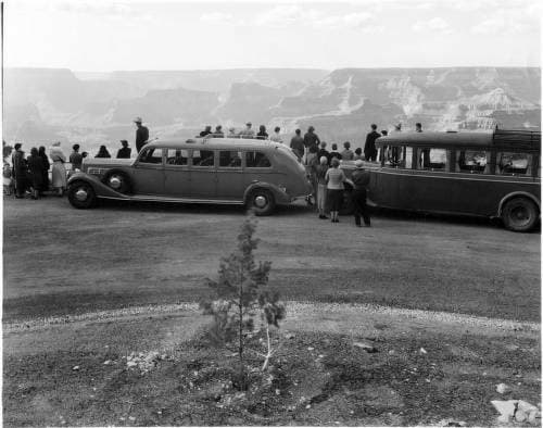 A black-and-white photo of two Harvey transportation cars that have let their passengers out to peer down at the Grand Canyon.There are probably about 40 men and women, all dressed in coats and hats, so it was likely chilly.