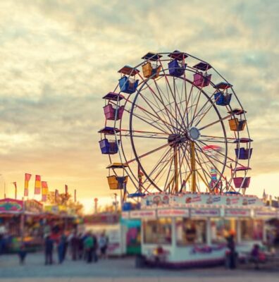 This is a color photograph of a modern ferris wheel with gondolas that probably hold 4 or 5 people. The photo shows some of the fair attractions and is set against the setting sun.