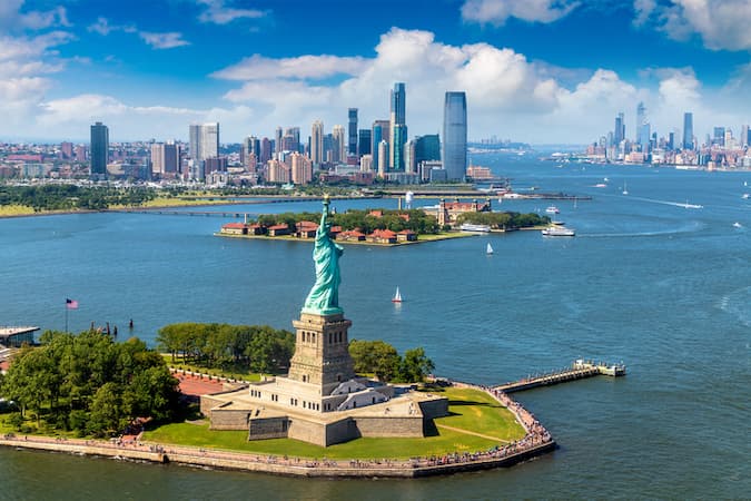 This is a color photograph of the New York harbor with the Statue of Liberty in the forefront of Ellis Island and the Manhattan skyline.