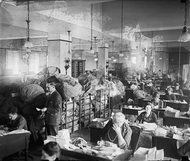 Black and white photo probably from about 1950 with women at desks working on letters and a man standing examining a letter in front of bags and bags of mail.