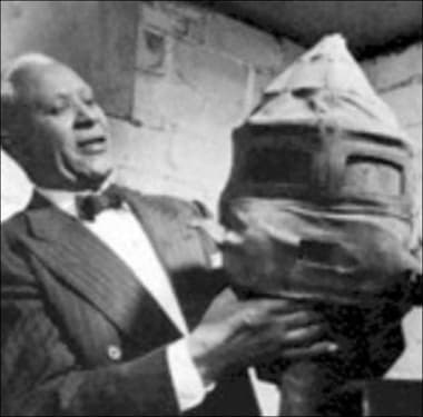 Black and white photograph of Garrett Morgan holding the gas mask he invented. It's large with pointed top and a 2 x 5 covered window for the wearer to look through