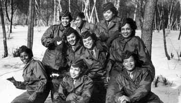 There is snow on the ground adn the women wear coats. They stand in front of a wooded area.