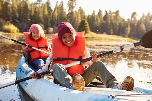 The kids are on a mountain lake in a boat that they are rowing together. Both have on life vests.