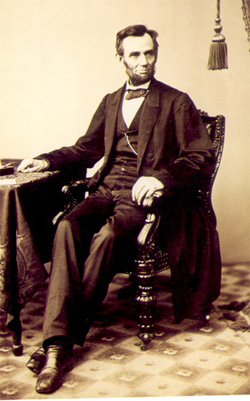 A sepia-toned photograph of Lincoln sitting beside a table. He looks off ot the right.