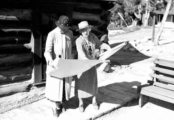 An older Mary Colter explains the blueprints for a buildlig to another woman who is with her. They are both dressed in hats and mid-calf dresses.