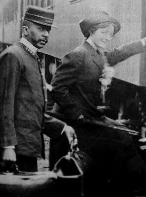 This is a black-and-white photo from the Library of Congress. A well-dressed woman is about to board a train. The Pullman porter stands just behind her waiting to carry on her bags. They both are looking at the camera.