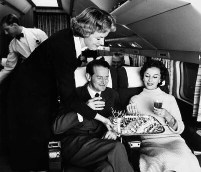 This may be a photo of first class on an early airplane. A waiter in the back wears a white coat and black bow tie. The passengers are elegantly dressed and the chairs look very comfortable. The flight attendant holds a try of snacks.
